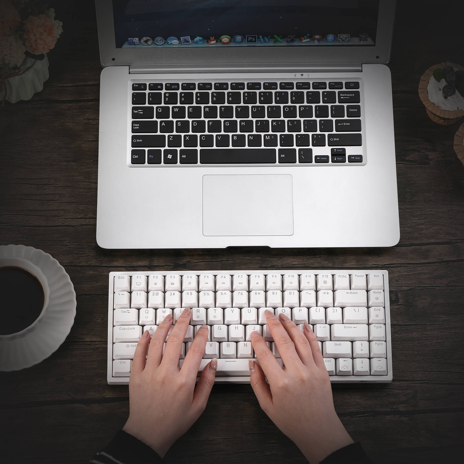  A user typing on a Royal Kludge RK84 compact gaming keyboard placed on a wooden table, alongside a laptop, a cup of coffee, and a decorative plate. The keyboard features a sleek white design and is positioned for easy access.